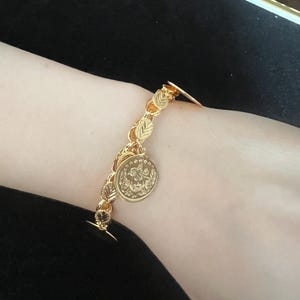 May include: A gold bracelet featuring a leaf-patterned chain and a circular coin pendant. The bracelet is fastened with a small clasp. The coin charm has an ornate design. The bracelet is worn on the wrist.