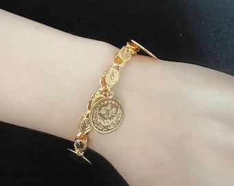 May include: A gold-colored bracelet with a leaf-like chain design and a round coin charm. The bracelet is secured with a small clasp and is worn on the wrist. The coin charm has an intricate design.