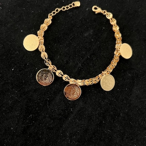 May include: Gold-toned bracelet with dangling coin charms. The bracelet features a delicate chain design with multiple circular charms. The charms have intricate patterns. The bracelet is displayed on a black surface.