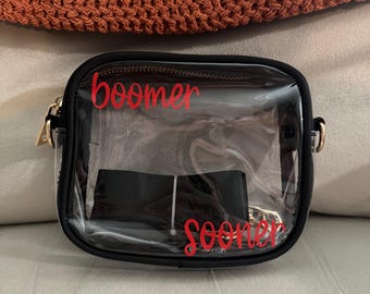 May include: A clear, rectangular crossbody bag with a black trim and strap. The bag has gold-colored zippers and the words "boomer sooner" in red script across the front.