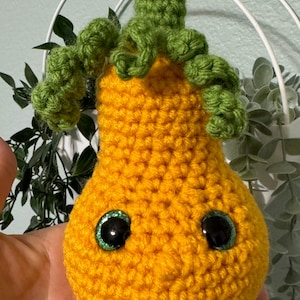 May include: A handmade, crocheted pear-shaped plush toy. The body is a vibrant yellow-orange, with green curly tendrils and a green stem. It has large, glittery black eyes. This whimsical item is likely a decorative piece or a child's toy.