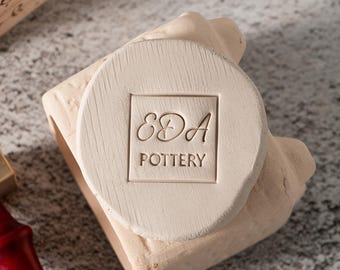 Puede incluir: Un sello de cerámica beige claro con el texto "EDA POTTERY" dentro de un cuadrado. El sello está sobre una forma de cerámica beige claro. El fondo es una superficie gris moteada.