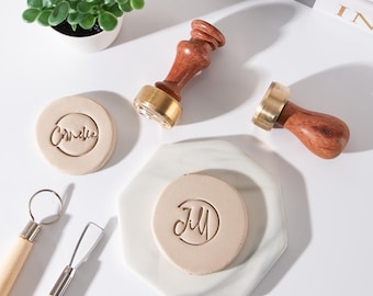 May include: Two wooden wax seal stamps with brass heads, one featuring the name Cornelia and the other Jill embossed on circular clay discs. Two metal sculpting tools with wooden handles are positioned nearby for crafting and detailing.