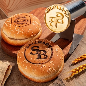 Op de afbeelding: Twee sesamzaad hamburgerbroodjes met aangepaste branding. Het bovenste broodje is gebrandmerkt met "Akis's Burgers" en het onderste broodje is gebrandmerkt met "SB BALLMARK".
