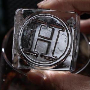 May include: A clear ice cube with the letter "H" embossed within a circular design. The ice cube is held in a person's hand, and the background is blurred, suggesting a drink or beverage.