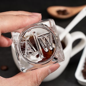 May include: A clear glass cube ice mold with the letters "AW" inside a circle. The ice mold is being held in a hand.