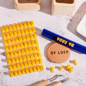 Puede incluir: Un conjunto de sellos de letras amarillas y herramientas para manualidades. Una herramienta azul con el texto "YOUJ YA" y un sello circular marrón con "BY LUCY" también están presentes. Las herramientas están sobre una superficie clara.