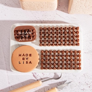Puede incluir: Una colección de sellos de galletas y sellos de letras marrones sobre una bandeja blanca. Un sello dice "MADE BY LISA" y otro "3DAM YA A21J". La bandeja también contiene juegos de sellos de letras individuales y una pequeña herramienta de metal.