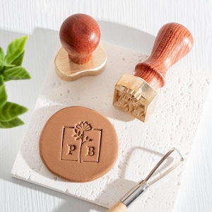 May include: A set of two brass stamps with wooden handles, one with a floral design and the letters "P" and "B" inside a square, and the other with a more intricate floral design. The stamps are on a white surface with a brown clay disc with the floral stamp impression.