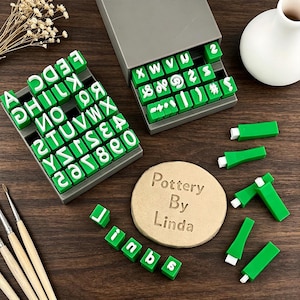 May include: A set of green and white rubber stamps featuring letters, numbers, and symbols arranged in gray storage trays. A circular tan clay disc is imprinted with the text Pottery By Linda, accompanied by several loose stamps and brushes.