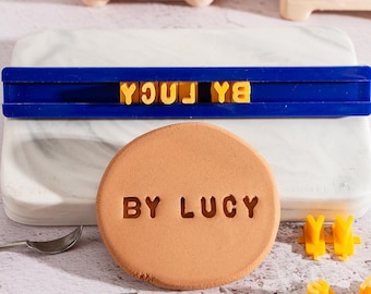 Puede incluir: Un sello de plástico azul con las letras "BY LUCY" en amarillo. El sello se utiliza para crear una impresión en un disco de arcilla de color terracota.
