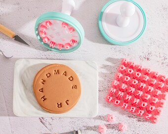 Puede incluir: Un sello para galletas circular de color verde menta con mango blanco, una base blanca a juego, una galleta redonda de color tostado con el texto HANDMADE JR grabado y una lámina de silicona rosa con letras individuales para personalizar.