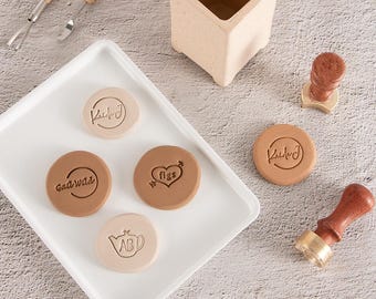 May include: A white rectangular tray holds four circular clay discs featuring embossed designs including the text "KukJ", "GadWild", "figs" with a heart, and a teapot with "ABD". Two wooden sculpting tools, a square clay container, and two wax stamps are present.