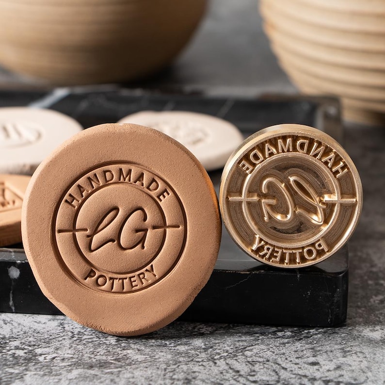 May include: Two round clay stamps, one with the text "HANDMADE LG POTTERY" and the other with the text "HANDMADE POTTERY" and a stylized "LG" logo. Both stamps are made of brass and have a circular design.