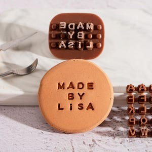 May include: A round, terracotta-colored stamp with the words "MADE BY LISA" embossed on it. A brown stamp with the reversed text "MADE BY YOU" is also visible. Additional letter stamps are also present.