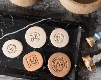 May include: Five circular and square clay pieces featuring various embossed logos including Linda Haymon Ceramics, Jill, Jane Wern Ceramics, AH Pottery, and LG Pottery, displayed on a black marble tray with two brass stamps with white handles.