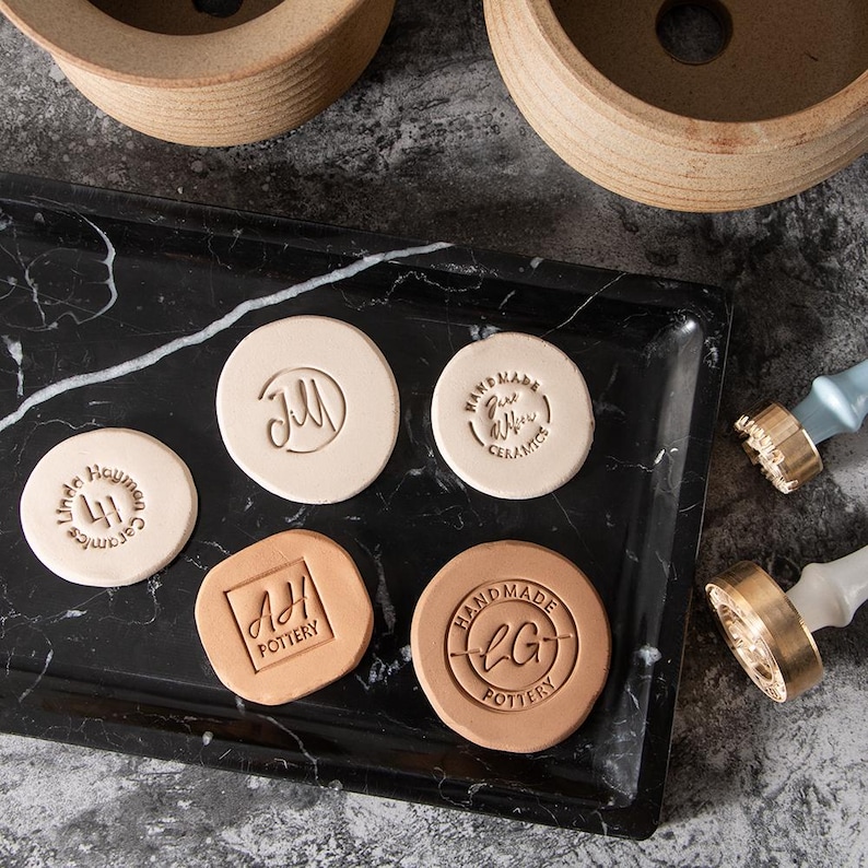 May include: Five different clay stamps with various designs and text. The stamps are on a black marble surface. The text on the stamps includes "Handmade", "Ceramics", "Pottery", "JM", "AH", and "LG".
