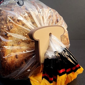 Puede incluir: Una hogaza de pan con semillas en una bolsa de plástico transparente, cerrada con un clip marrón con forma de pan. El clip se inserta a través de la abertura de la bolsa. El pan tiene una corteza dorada y semillas visibles.