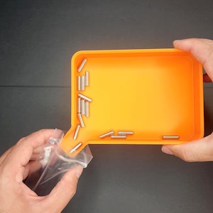 May include: An orange rectangular tray with a spout, filled with small, silver cylindrical objects. A clear plastic bag is being emptied into the tray. The background is a dark grey surface.