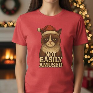 May include: Red t-shirt featuring a grumpy cat wearing a Santa hat. The cat has brown and white fur with a stern expression. The text "NOT EASILY AMUSED" is printed below the cat in a vintage style font.
