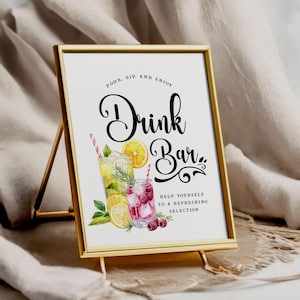 May include: A framed sign with the words "Drink Bar" in elegant script. The sign features watercolor illustrations of refreshing drinks with lemons and raspberries. The sign is in a gold metal frame and is displayed on a beige surface.