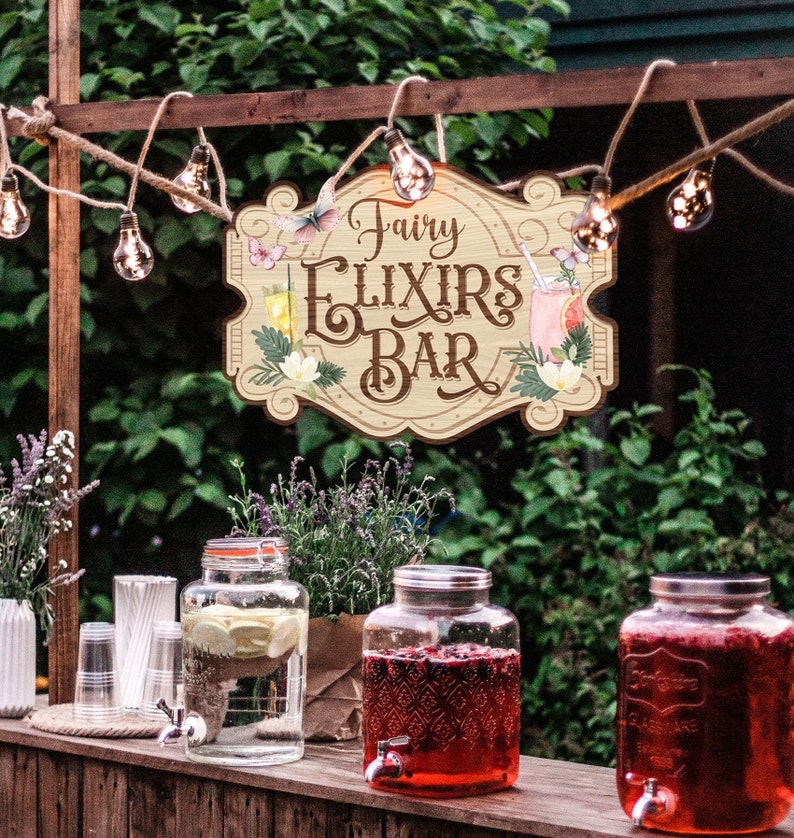 K&ouml;nnte beinhalten: Ein Holzschild mit dem Text "Fairy Elixirs Bar" in einer verspielten Schriftart. Das Schild ist mit Blumen, Schmetterlingen und einem Glas Limonade verziert. Zwei Glasbeh&auml;lter mit Ausgie&szlig;ern sind mit roter und klarer Fl&uuml;ssigkeit gef&uuml;llt und stehen auf einer Holztheke.