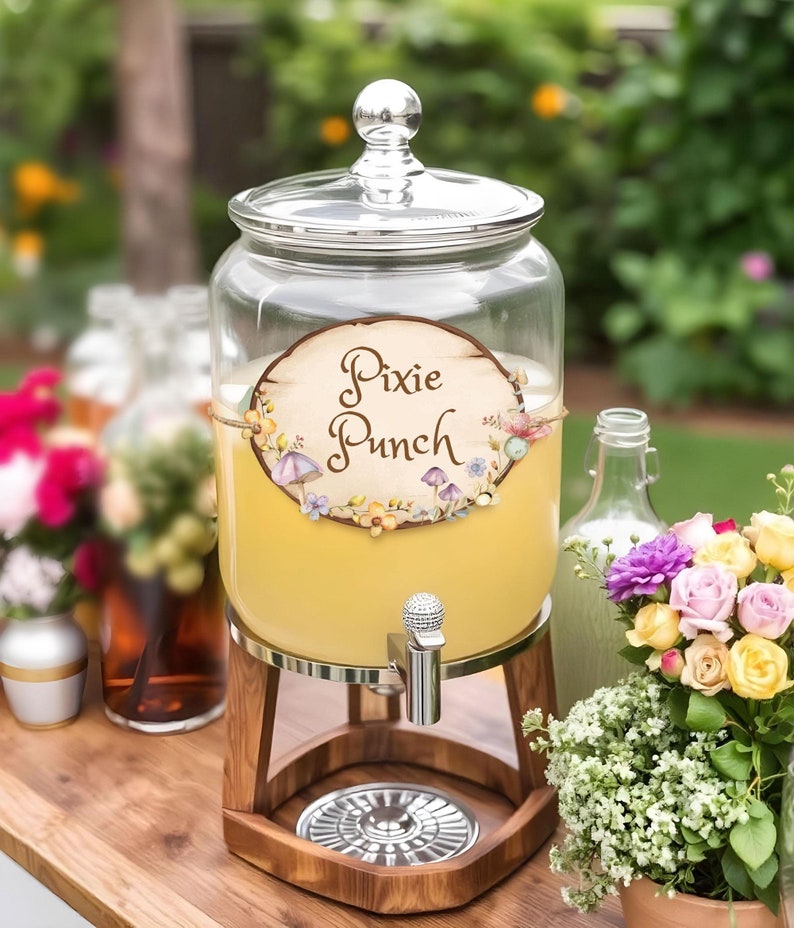 May include: A clear glass beverage dispenser with a silver spigot and wooden stand. The dispenser is filled with yellow liquid and has a label that reads "Pixie Punch". The label is decorated with flowers and mushrooms. A glass lid sits on top.