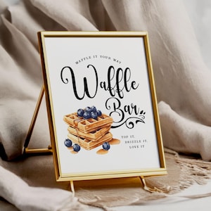 Peut inclure: Un panneau doré avec l'inscription "Waffle Bar" en noir. Le panneau présente une illustration à l'aquarelle de gaufres garnies de myrtilles et de sirop. Le texte "Top it, Drizzle it, Love it" est également inclus.
