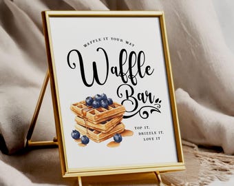 May include: A gold-framed sign with the words "Waffle Bar" in elegant black script. The sign features a watercolor illustration of waffles topped with blueberries and syrup. The text "Top it, Drizzle it, Love it" is also included.