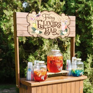 K&ouml;nnte beinhalten: Ein Getr&auml;nkewagen aus Holz mit einem Schild, auf dem "Fairy Elixirs Bar" steht. Es gibt drei Glasbeh&auml;lter, die mit bunten Fl&uuml;ssigkeiten gef&uuml;llt sind, und Plastikbecher.