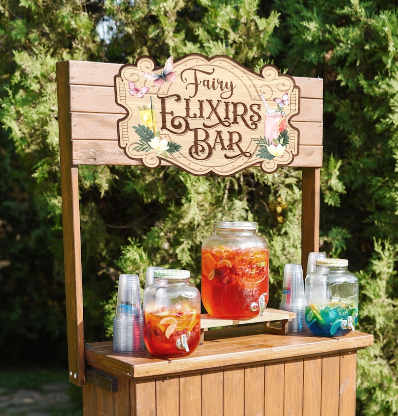 K&ouml;nnte beinhalten: Ein Getr&auml;nkewagen aus Holz mit einem Schild, auf dem "Fairy Elixirs Bar" steht. Es gibt drei Glasbeh&auml;lter, die mit bunten Fl&uuml;ssigkeiten gef&uuml;llt sind, und Plastikbecher.