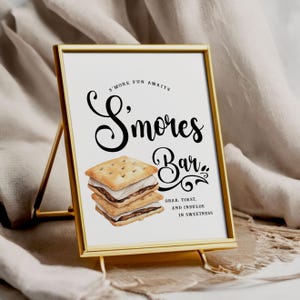 May include: A framed sign with a gold metal stand. The sign reads "S'mores Bar" in elegant script, with an illustration of s'mores. The text "Grab, Toast, and Indulge in Sweetness" is also included. The background is white.