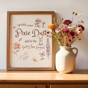 May include: A framed print with a white background and brown text that reads "Sprinkle some Pixie Dust and let the fairy fun begin!" The print features a watercolour illustration of a bunny rabbit, mushrooms, butterflies, and flowers.