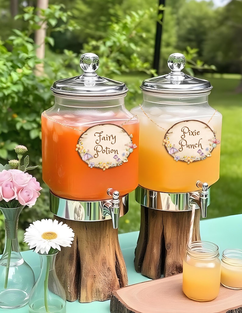 May include: Two glass beverage dispensers filled with orange and yellow liquid, labeled "Fairy Potion" and "Pixie Punch." Each dispenser has a silver lid and spigot, and sits on a wooden base. A vase of pink roses and a white daisy are in the foreground.
