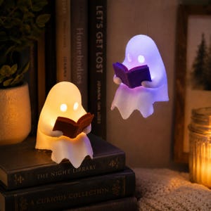 May include: Two ghost-shaped LED lamps, one sitting on a stack of books and one suspended in the air. The sitting ghost glows with a warm yellow light and holds a brown book. The floating ghost glows with a purple light and holds a purple book.