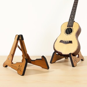 May include: Two wooden ukulele stands with personalized engravings. The stand on the left has the text "Eat. Drink. & Make Music" and the stand on the right has the text "Rock On".