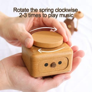May include: A wooden music box with a round, wooden knob on top. The text "Rotate the spring clockwise 2-3 times to play music" is printed on the top of the box.