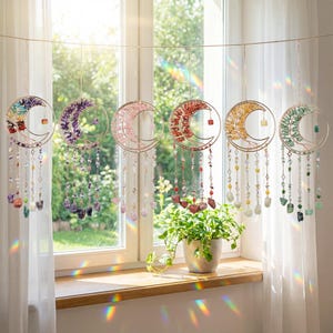 May include: Six crescent moon suncatchers with gemstone fillings and beaded strands, suspended in front of a window. Each has a gold-coloured metal frame. Gemstones are in shades of purple, pink, red, yellow, and green. The window lets in sunlight.