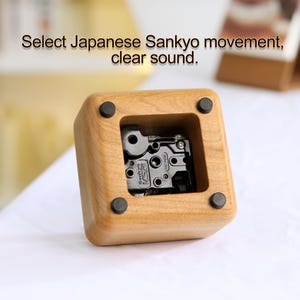 May include: A wooden music box with a clear plastic window revealing a black Sankyo music movement. The music box is made of light brown wood and has a square shape.