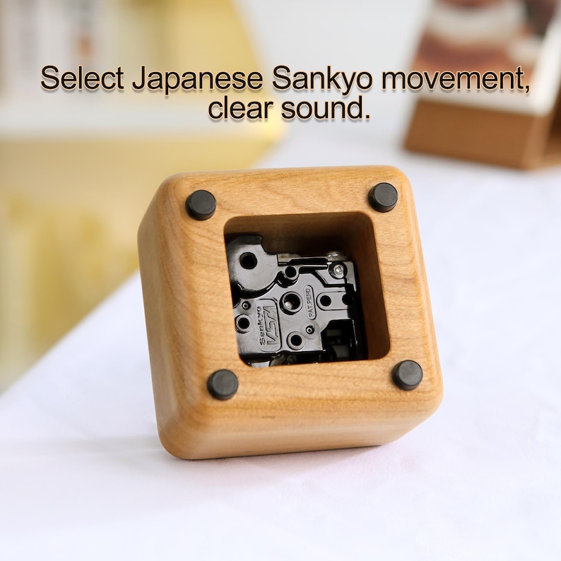 May include: A wooden music box with a clear plastic window revealing a black Sankyo music movement. The music box is made of light brown wood and has a square shape.