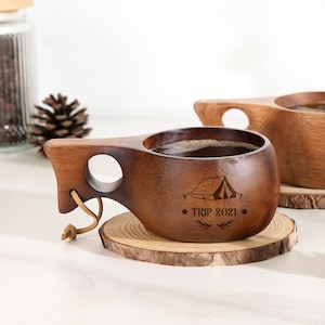 Op de afbeelding: Een houten campingmok met een handvat en een leren riem. De mok heeft een donkerbruine afwerking en heeft een tentafbeelding met de tekst "TRIP 2021". De mok staat op een houten onderzetter.