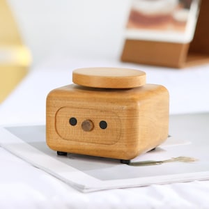 May include: A wooden music box with a round top and a small knob on the front. The box is light brown and has a simple design.