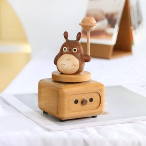 May include: A wooden music box with a brown and white cartoon character holding a star. The music box is made of light wood and has a crank on the side.