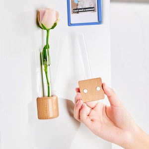 May include: A magnetic vase with a wooden base and a glass tube for holding a single pink rose. The vase is attached to a white surface with a magnetic backing. The text "I just need a moment" is visible on a blue photo frame in the background.