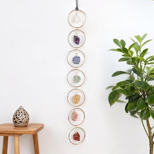 May include: A decorative wall hanging with seven gold-tone metal rings, each displaying a different coloured raw crystal. The crystals are clear, purple, blue, green, yellow, pink, and red. A black cord suspends the rings, creating a natural aesthetic.
