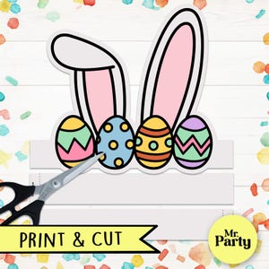 May include: Printable Easter bunny ears headband with a white background and pink ears. The headband is decorated with four colorful Easter eggs with different patterns. The text "PRINT & CUT" is at the bottom of the image.