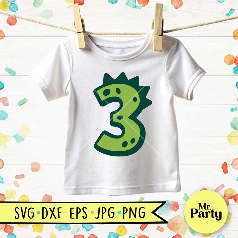 Pu&ograve; includere: Maglietta bianca per bambini con un design verde del numero 3. Il numero &egrave; disegnato per assomigliare a un dinosauro, con una parte superiore a spillo e dettagli a pois. La maglietta &egrave; appesa a un filo per stendere.