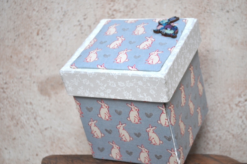 Op de afbeelding: Een kleine, vierkante, blauwe en witte geschenkdoos met een patroon van witte konijnen. De deksel van de doos heeft een wit bloemenpatroon en een houten konijnendecoratie.