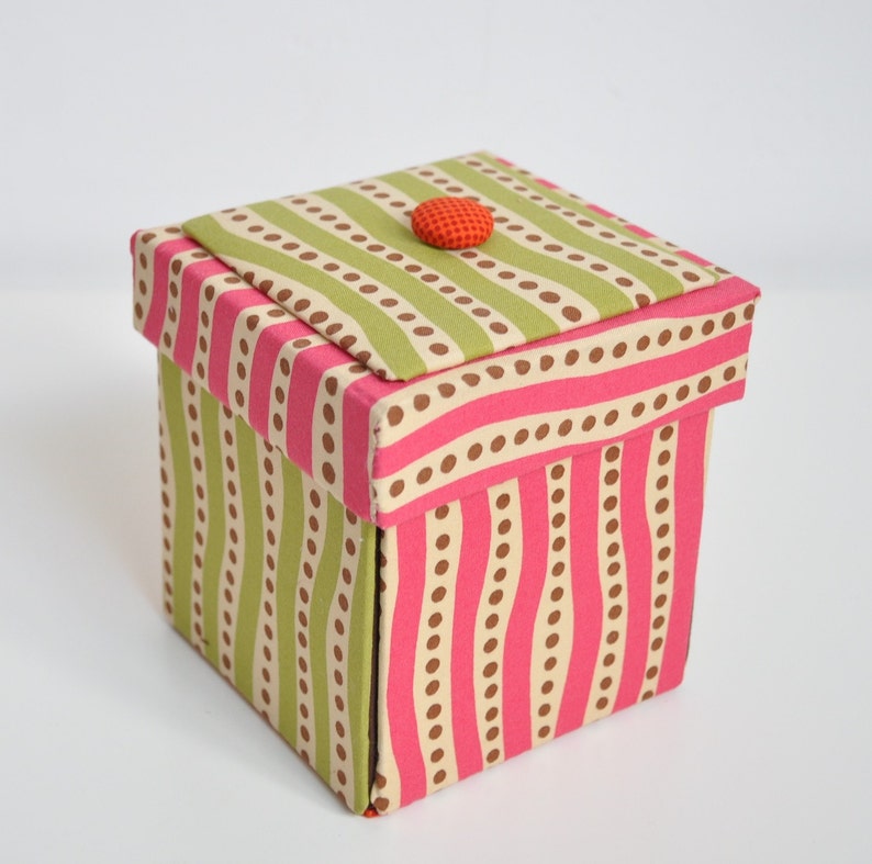 Op de afbeelding: Een kleine, vierkante doos met een roze en groene gestreepte print en bruine stippen. Het deksel is gesloten en heeft een enkele oranje knop bovenop.