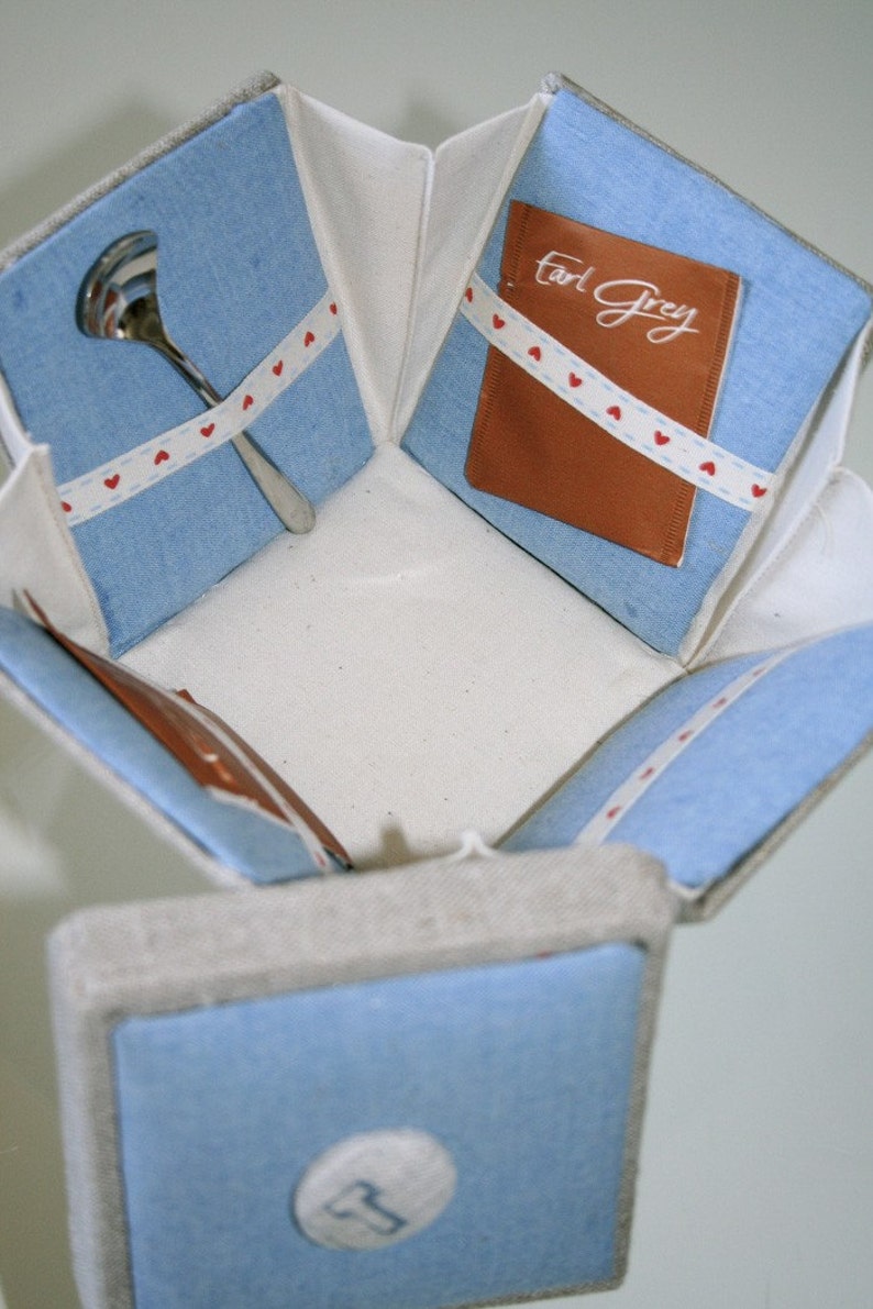 Op de afbeelding: Een blauwe en witte stoffen doos met een wit interieur. De doos bevat een klein lepeltje en een theezakje. Het theezakje is gelabeld met "Earl Grey".